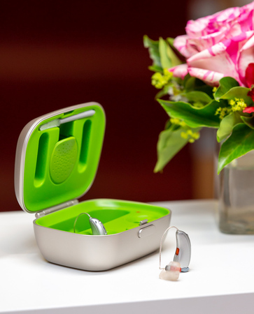 Modernes Hörgerät mit Ladestation auf Tisch, daneben ein Blumenstrauß, Hörakustik.