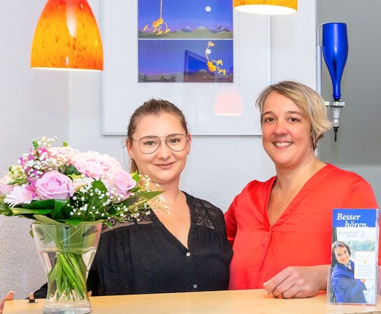 Zwei Frauen lächeln an einem Empfangstresen mit Blumenstrauß und Hörakustik-Flyer.